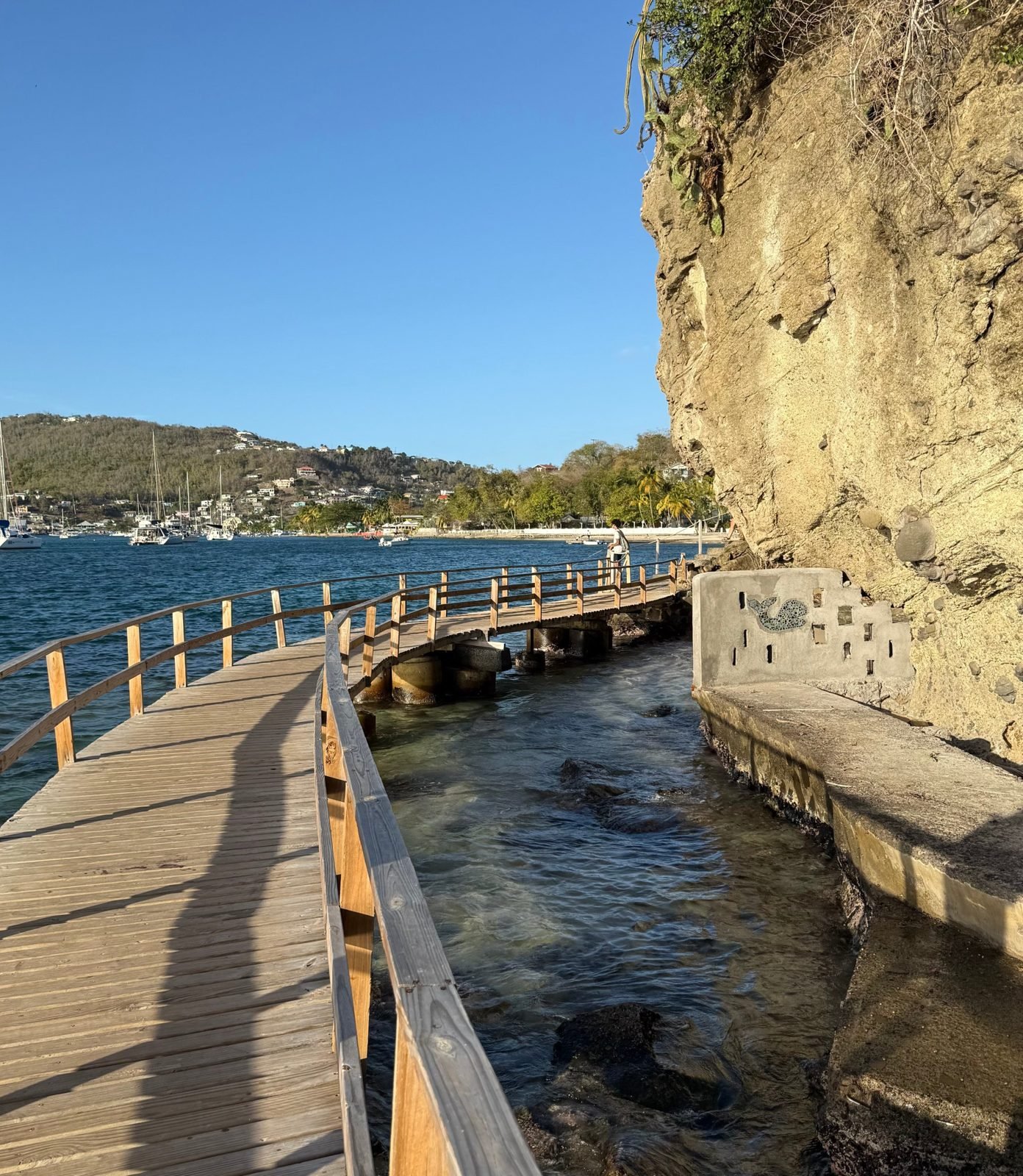 Waterfront boardwalk along a cliff, with sailboats in the bay